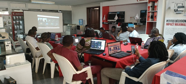 Group of people seated around a table with laptops, a mouse, bottles, and other items, participating in a digital marketing workshop; banner with text, display screen, door, and ceiling lights visible in the background.