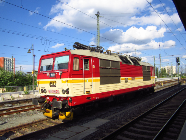 Rote und gelbe Elektrolokomotive auf Schienen neben einem Bahnsteig, mit Strommasten, Laternen, Bäumen, Gebäuden und einem bewölktem Himmel im Hintergrund.