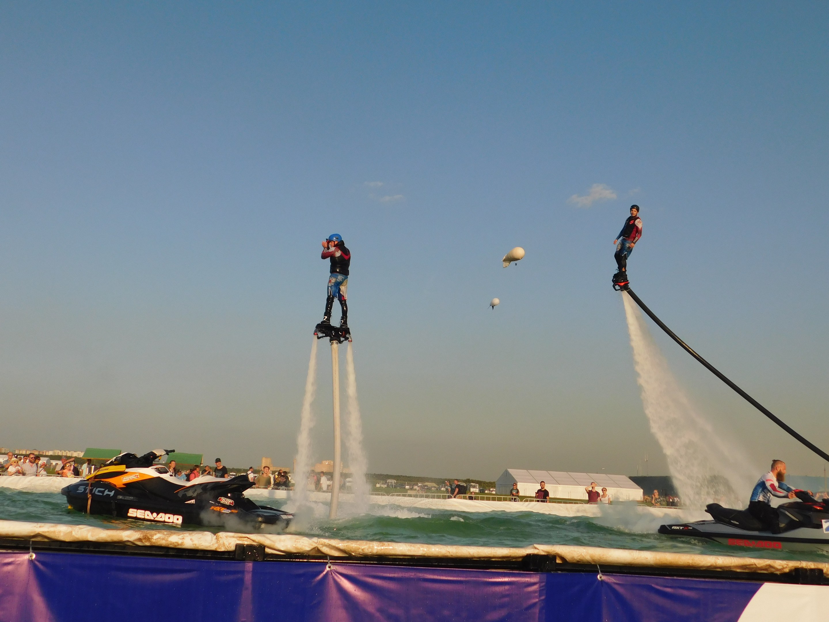 Group of people riding jet skis over water with a banner below, set against a backdrop of buildings, people, and a clear blue sky.