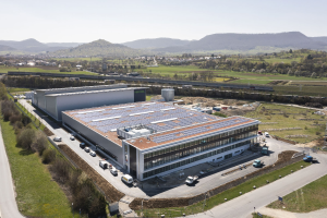 Aerial view of a large building with solar panels on its roof, surrounded by vehicles, trees, grass, and poles, set against hills and a clear blue sky, identified as Germany's first solar-powered factory.