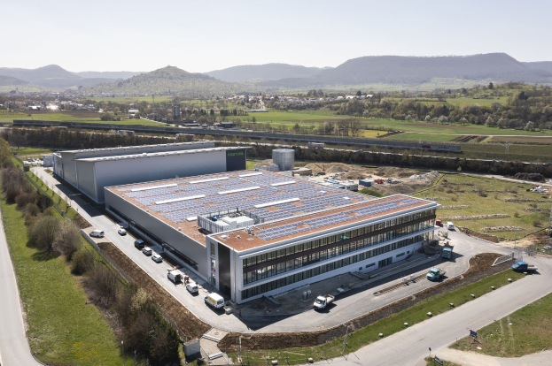 Aerial view of a large building with solar panels on its roof, surrounded by vehicles, trees, grass, and poles, set against hills and a clear blue sky, identified as Germany's first solar-powered factory.