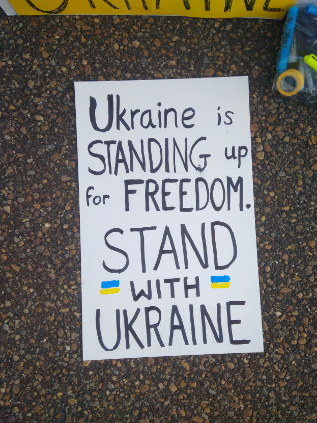Ein weißer Zettel mit den Worten "Ukraine steht für Freiheit, steht mit Ukraine" liegt vor einer gelben Fahne auf dem Boden, rechts daneben eine Plastikabdeckung mit verschiedenen Gegenständen.
