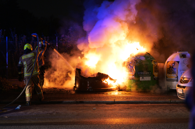 Ein Feuerwehrmann in Helm und Ausrüstung steht mit einer Rohrleitung vor einem brennenden Auto an der Straße, mit anderen Fahrzeugen rechts im Bild, einem Zaun und Bäumen im Hintergrund und einem dunklen Himmel.