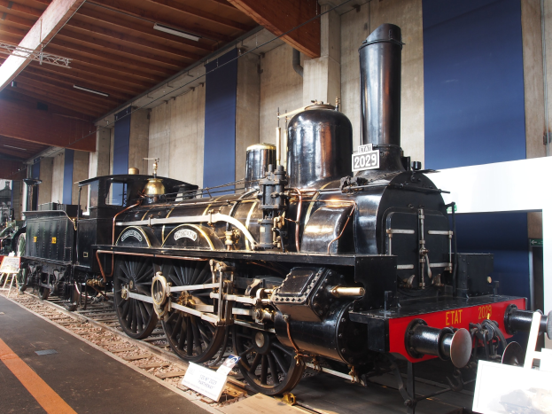 Alte Dampflokomotive in einem Museum ausgestellt, umgeben von Eisenbahnschienen und Informationsschildern, mit einer Wand und beleuchteter Decke im Hintergrund.