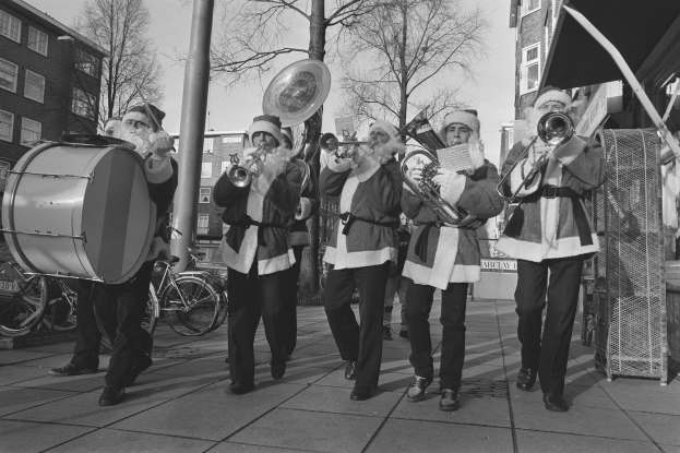 Eine Gruppe von Männern in Santa Claus-Kostümen, die auf einem Bürgersteig Musikinstrumente spielen, umgeben von Fahrrädern, Pfählen, Bäumen, Gebäuden mit Fenstern, unter einem klaren Himmel, dargestellt in Schwarz-Weiß.