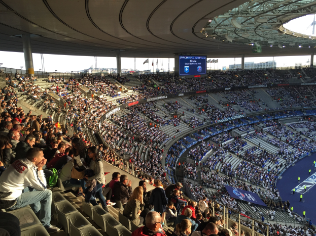 Große Menschenmenge im Allianz Arena Stadion bei einem Füßtballspiel, mit einer Bühne auf der rechten Seite, Fahnen, Masten, einem Bildschirm und sichtbarem Himmel im Hintergrund.