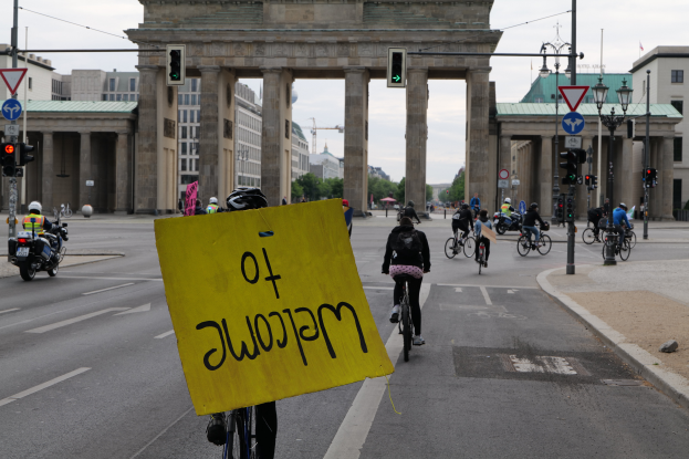 Eine Gruppe von Menschen auf Fahrrädern fährt auf der Straße vor dem Reichstag in Berlin, Deutschland, mit Helmen, wobei eine Person ein gelbes Schild hölt, umgeben von Laternenmästen, Verkehrszeichen, Gebäuden, Bäumen und einem klaren blauen Himmel.