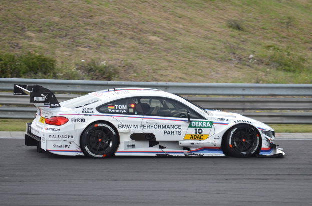 BMW M4 DTM-Rennwagen auf einer Rennstrecke mit Text auf dem Fahrzeug, umgeben von grünem Gras und einer Begrenzung im Hintergrund.