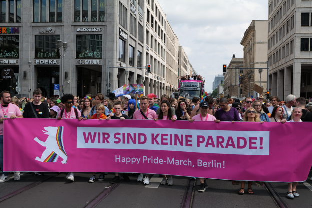 Eine Gruppe von Menschen marschiert auf einer Straße in Berlin mit einer pinken "Happy Pride March"-Fahne, mit Gebäuden, Laternenmasten und Verkehrszeichen an der Straße unter einem bewölkten Himmel.