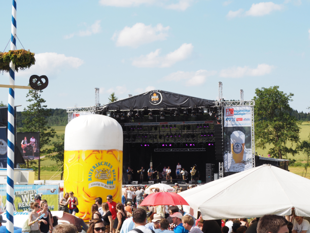 Eine große Menschenmenge beim Oktoberfest in München, Deutschland, viele tragen Kappen und Goggles, einige halten Schirme, im Vordergrund ein Zelt, links ein dekorierter Pfahl, im Hintergrund Bäume und ein bewölkter Himmel sowie eine Bühne mit einem Banner und Musikern.