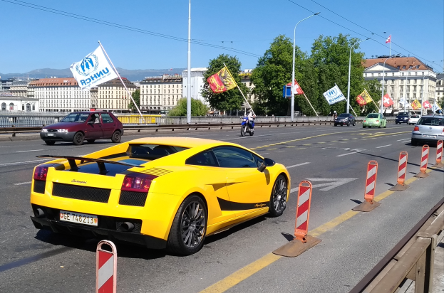 Ein gelber Sportwagen fährt auf einer Straße neben einer Brücke, umgeben von Verkehrskegeln, Laternenmasten, Fahnen, Bäumen, Gebäuden und einem Geländer, mit Hügeln und einem klaren blauen Himmel im Hintergrund.