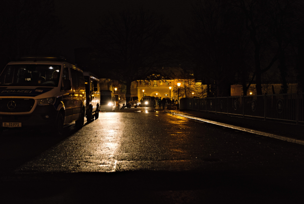 Ein Polizeiwagen steht nachts am Straßenrand, mit Fahrzeugen auf der Straße, einem Zaun rechts, Bäumen, Laternenmasten und einem Gebäude im Hintergrund.
