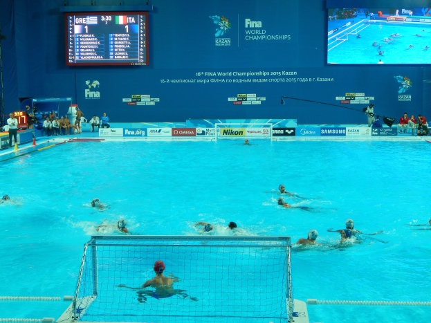 Wasserballspieler bei den FINA-Weltmeisterschaften 2016 in Kazan im Pool gegeneinander antretend, mit Zuschauern auf der linken Seite und einem Anzeigeboard mit Event-Branding im Hintergrund.