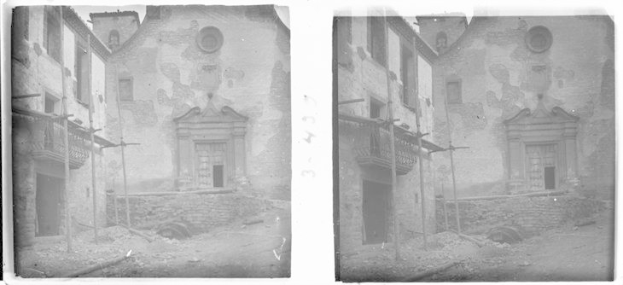 Black and white photo of a dilapidated building with crumbling walls, broken windows, and doors, surrounded by poles and other objects.