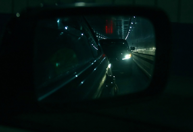 Ein Auto fährt durch einen Tunnel in der Nacht, wobei sein Seitenspiegel andere Fahrzeuge reflektiert, die von Tunnel- und Autolichtern beleuchtet werden.