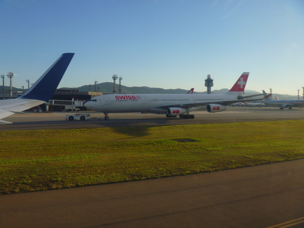 Schweizer International Air Lines Boeing 777-300ER auf der Rollbahn am San Francisco International Airport mit umgebenem Gras, einer Straße, Laternenmasten, Gebäuden, Hügeln und einem klaren blauen Himmel.
