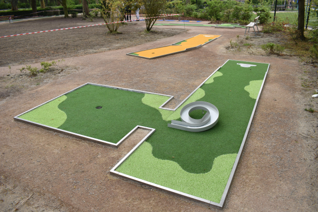 Ein Minigolfplatz in einem Park, umgeben von grünem Gras, Pflanzen und Bäumen, mit Menschen in der Nähe und Gebäuden im Hintergrund unter einem klaren blauen Himmel.