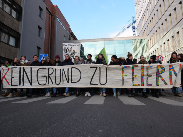 Demonstranten auf einer Straße halten ein Transparent mit der Aufschrift "Kein Grund zu Feiern" gegen die deutschen Sparmaßnahmen, mit Gebäuden, einem Schild, einem weiteren Text-Transparent, einem Kran und einem bewölkten Himmel im Hintergrund.