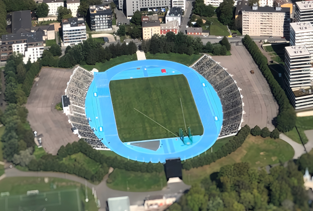 Aerial view of the Olympic Stadium in Munich, Germany, surrounded by city infrastructure including buildings, trees, roads, vehicles, and grass.