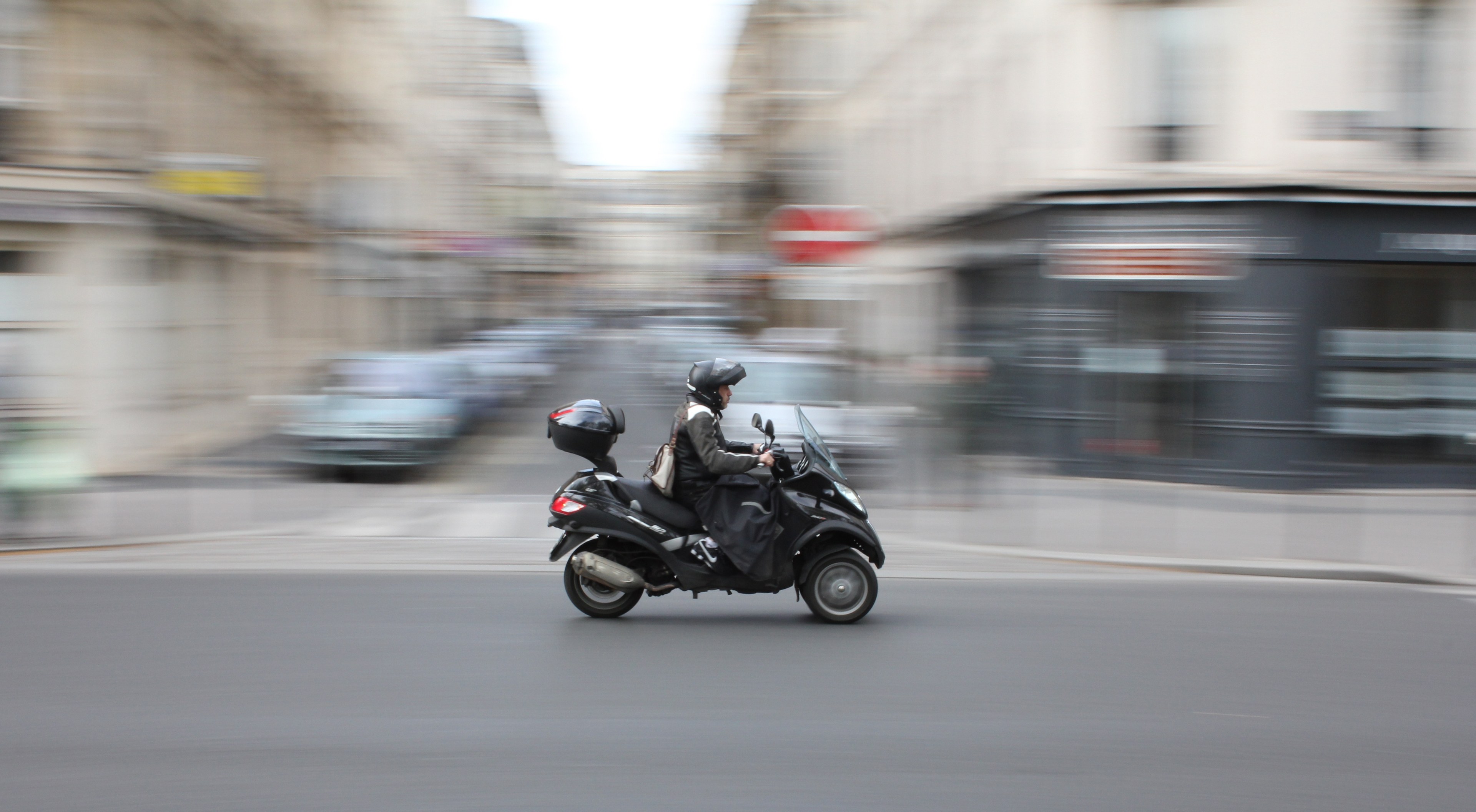 Eine Person in schwarzer Kleidung und Helm fährt auf einem Motorroller auf einer Stadtstraße mit anderen Fahrzeugen, Gebäuden und einem klaren blauen Himmel im Hintergrund.