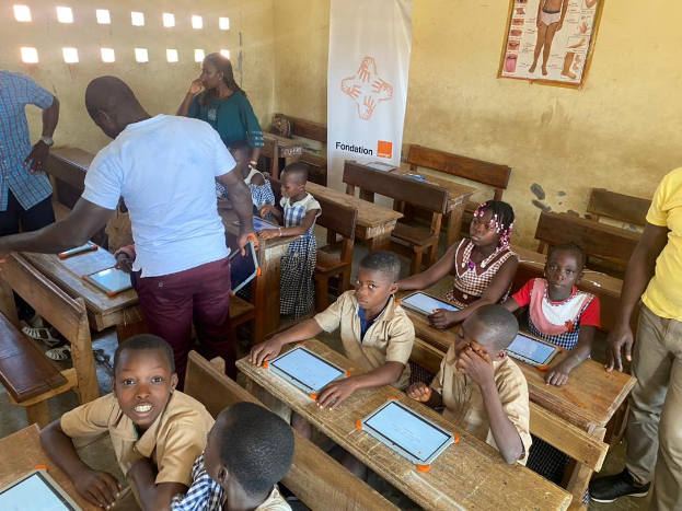 Eine Gruppe von Kindern sitzt an Tischen in einem Klassenzimmer, nutzt Tablets, mit einigen Menschen drumherum und einem Banner und Plakat im Hintergrund.