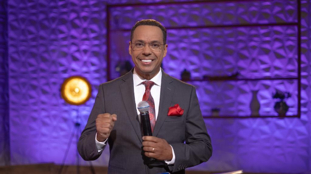 Stephen Colbert, Moderator von *The Daily Show with Trevor Noah*, steht auf einer Bühne und hält ein Mikrofon, trägt einen Anzug, eine Krawatte und eine Brille, während im Hintergrund ein Scheinwerfer und ein Ständer zu sehen sind.