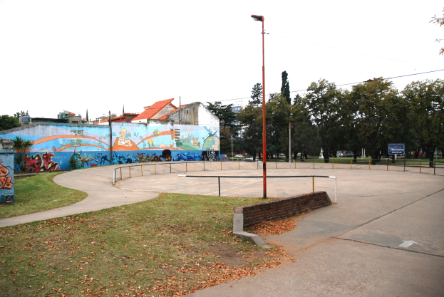 Skate park with a mural on a building, surrounded by grass, dried leaves, light poles, railings, and a painted wall, set against a backdrop of trees, buildings, vehicles, and sky.