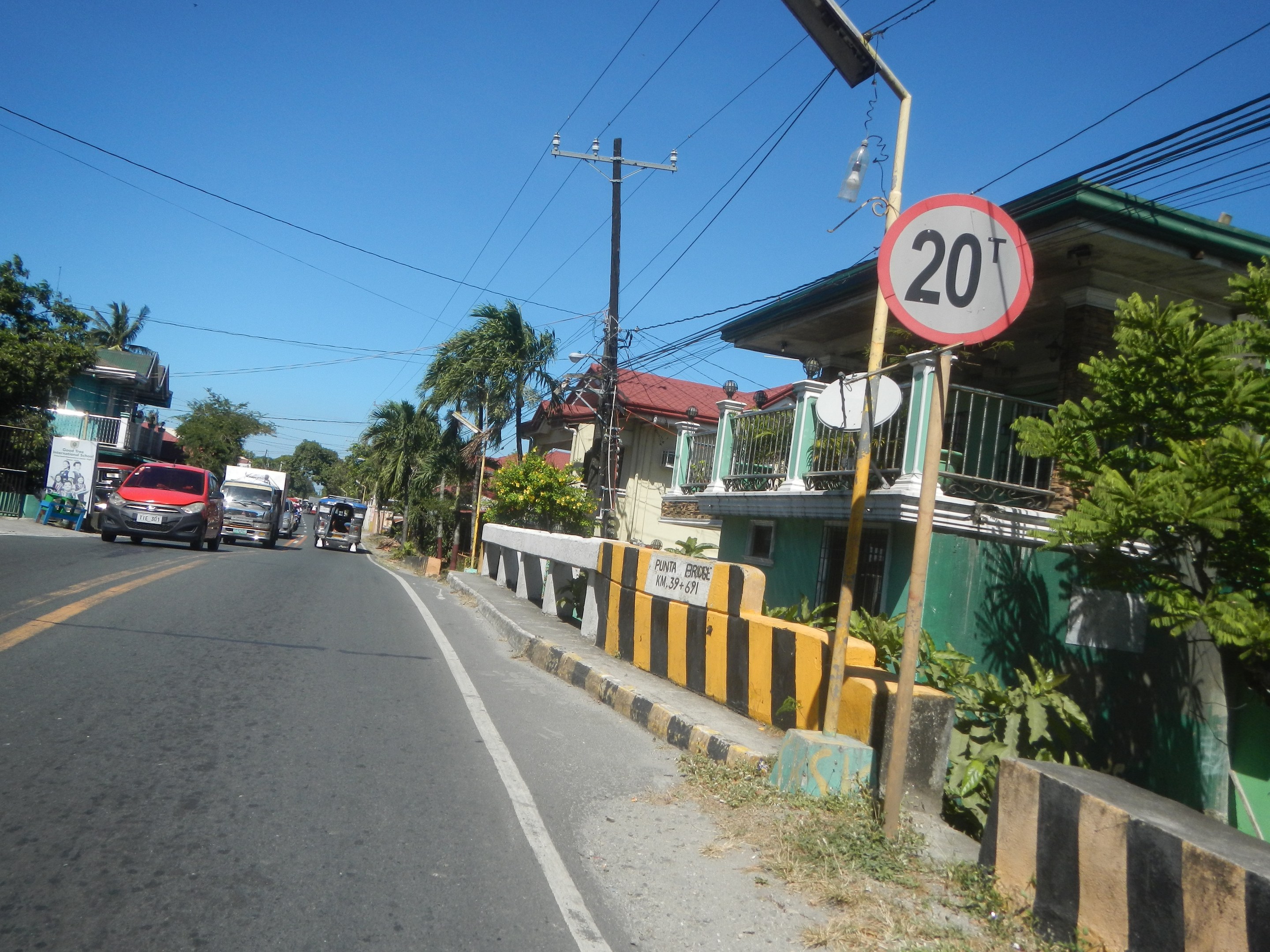 A street with moving cars, a speed limit sign, electric poles with wires, buildings with windows and railings, trees, and a clear blue sky.