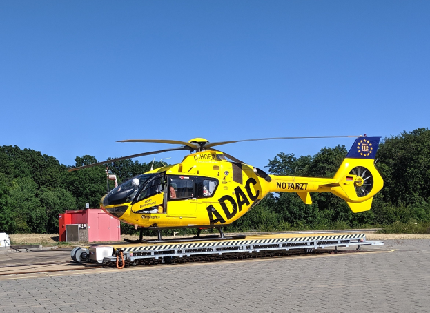 Ein gelber Hubschrauber steht auf einem Flughafen-Asphalt umgeben von Bäumen und einem klaren blauen Himmel, mit verschiedenen Gegenständen rund um den Hubschrauber verstreut.