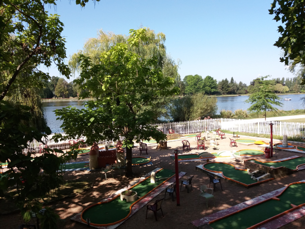 Ein Minigolf-Platz in einem Park mit grünem Gras, Bäumen und einem Zaun, mit Stühlen und Tischen, einem Gewässer mit einem Boot und einem sichtbaren Himmel.