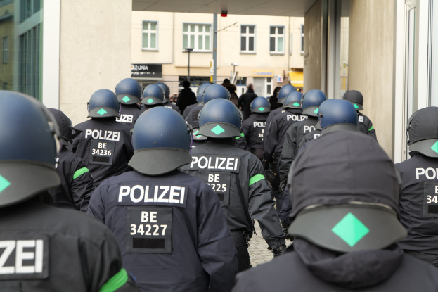 Eine Gruppe von Polizisten in Uniform und Helmen vor einem Gebäude stehend, mit Menschen, Pfählen und anderen Gebäuden im Hintergrund.