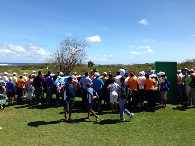 Eine Gruppe von Menschen auf einem grünen Feld bei einem Golfturnier, einige tragen Mützen und tragen Taschen, mit Bäumen, Pflanzen, einer bewölkten Himmel und einem Gewässer im Hintergrund.