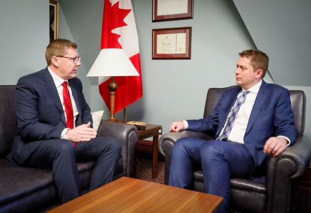 Zwei Männer im Blazer und Krawatte mit Brille vor einer kanadischen Flagge, mit einer Lampe und anderen Gegenständen auf dem Tisch dazwischen und gerahmten Gegenständen an der Wand dahinter.