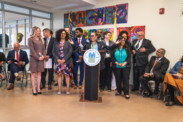Gruppe von Menschen in einem Raum mit einem Podium, einem Mikrofon und einer Tafel mit dem Text "Sancho City Councilman Joins City Council", umgeben von Flaggen, Stühlen und einem Feuerlöscher.