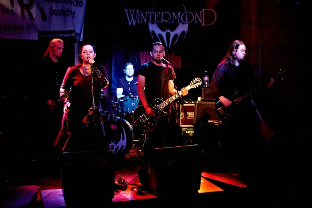 Eine Gruppe von Menschen, die auf einer Bühne mit Gitarren, Mikrofonen, einem zentralen Lautsprecher und einem Textbanner in einer dunklen, mysteriösen Umgebung spielen.