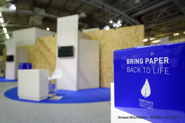 Messestände mit einem blauen und weißen "Bring Paper Back to Life"-Schild auf der rechten Seite, mit unscharfen Stühlen, Tischen, einem wandmontierten Fernseher und Deckenleuchten im Hintergrund.