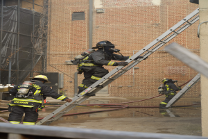 Eine Gruppe von Feuerwehrleuten in Helmen und Zylindern klettert an einer Leiter vor einem Backsteingebäude hoch, mit Rohren auf dem Boden und einer Metallstange unten und einem Gebäude mit Fenstern und einem Netz auf der linken Seite im Hintergrund.