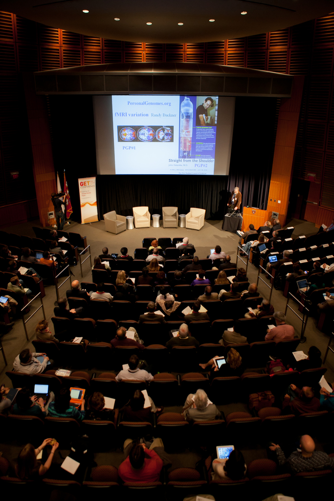 Ein Auditorium mit vielen sitzenden Menschen, die Gegenstände halten, zahlreichen Lichtern, Fahnen auf der linken Seite, einem Podium auf der rechten Seite und einer Werbetafel auf der linken Seite.