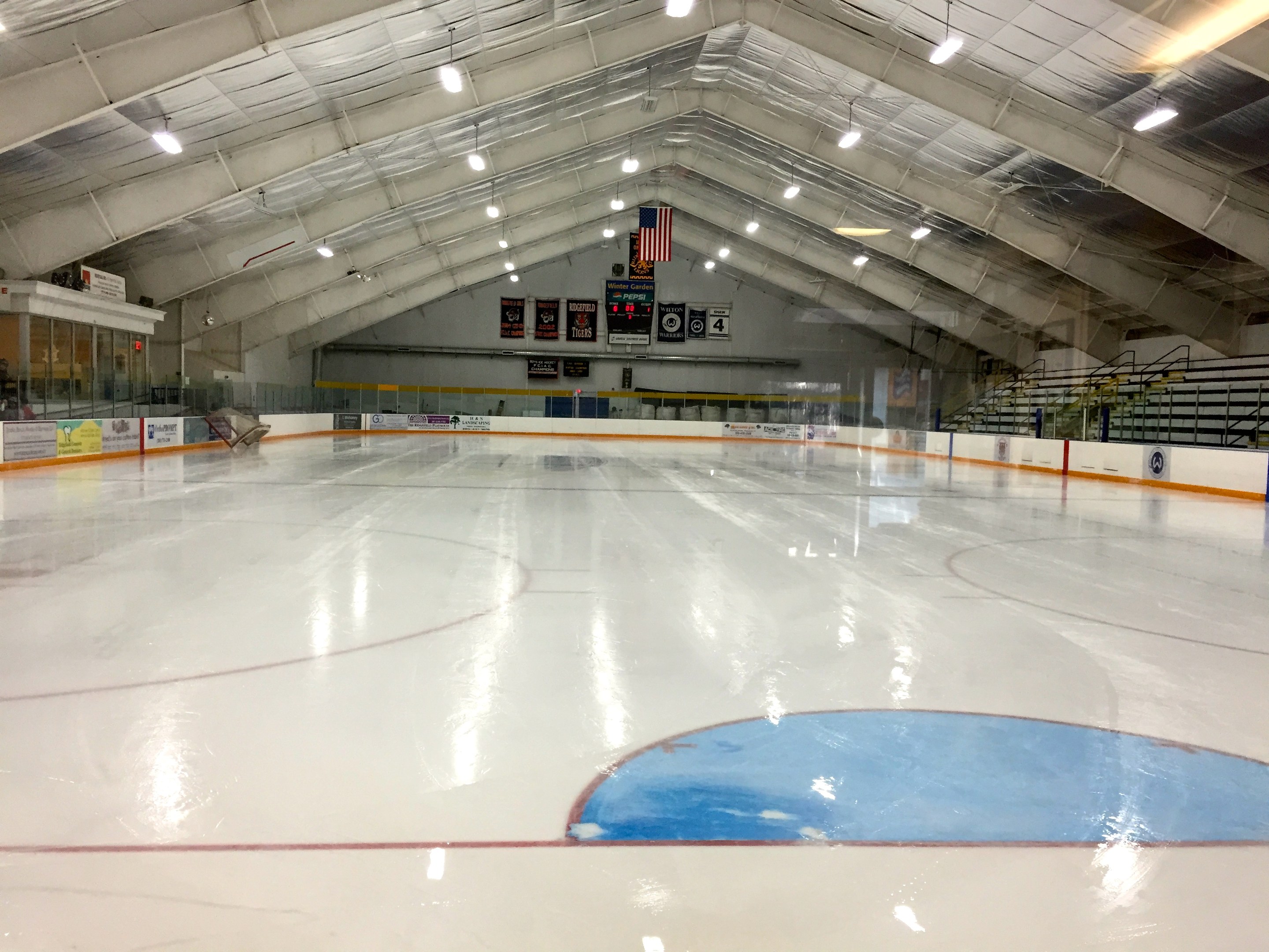 Große Indoor-Eisfläche mit Deckenbeleuchtung, Werbetafeln, einer Rückwand, Treppen mit Geländern und einigen Menschen, die einem Hockey-Sportplatz ähnelt.