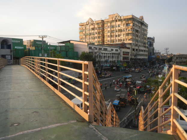 Eine Fußgängerbrücke mit Geländern überspannt eine vielbefahrene Straße mit Fahrzeugen und Fußgängern, im Hintergrund sind Gebäude, Bäume und Strommasten unter einem bewölkten Himmel zu sehen.
