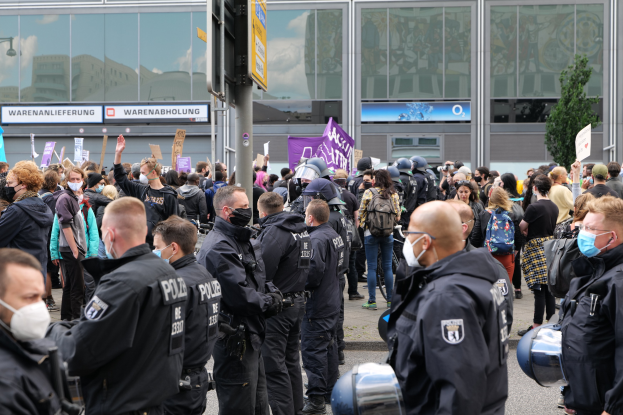 A large group of people protesting in front of a building, some holding placards and wearing helmets, with a signpost in the foreground and a tree in the background.