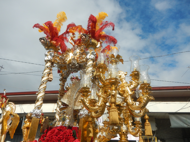 Ein großer goldener und roter Festwagen, der mit Blumen und anderen Dekorationsartikeln geschmückt ist, bei einem Karnevalsumzug, mit einem Gebäude, Strommasten mit Drähten und einem bewölkten Himmel im Hintergrund.
