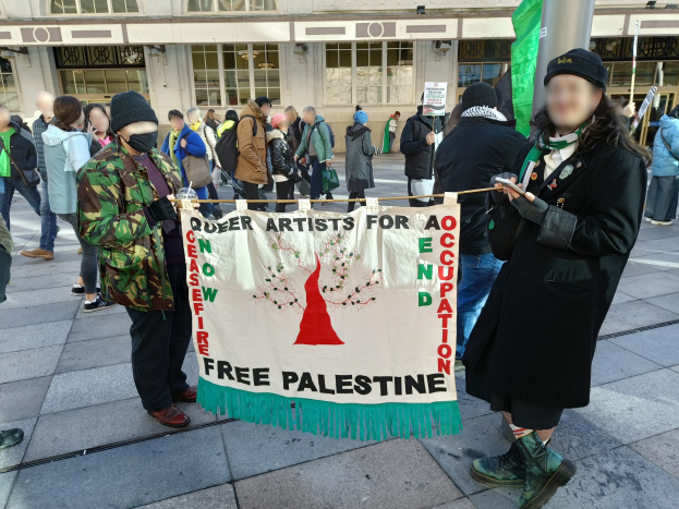 Gruppe von Menschen mit einer "Free Palestine"-Fahne vor einem Gebäude mit Fenstern und einer Tafel, mit einem Pfahl im Hintergrund.