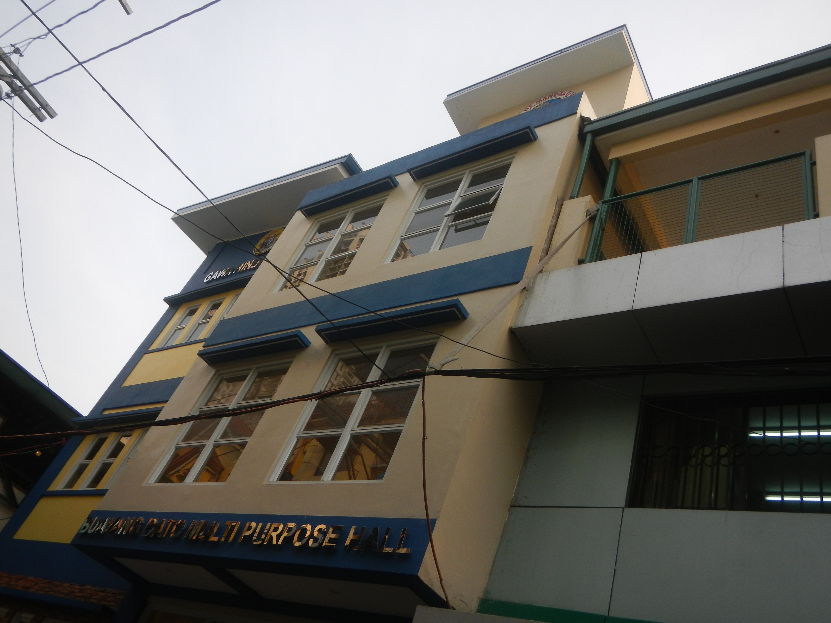 Mehrzweckgeb├Ąude mit blauer und gelber Fassade, Glasfenstern, einem Schild, einem Strommast mit Kabeln und einem klaren blauen Himmel im Hintergrund.