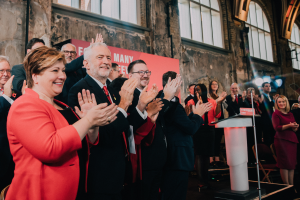 Eine Gruppe von Menschen, wahrscheinlich Liberale, klatscht feierlich vor einer Menge, mit einem Podium, Mikrofon und Texttafel rechts, sowie Stühlen, einer Fahne, Wand, Fenstern und Lichtern im Hintergrund.