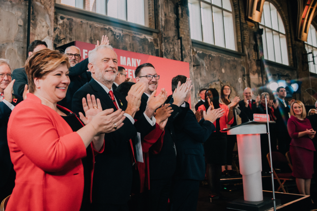Eine Gruppe von Menschen, wahrscheinlich Liberale, klatscht feierlich vor einer Menge, mit einem Podium, Mikrofon und Texttafel rechts, sowie Stühlen, einer Fahne, Wand, Fenstern und Lichtern im Hintergrund.