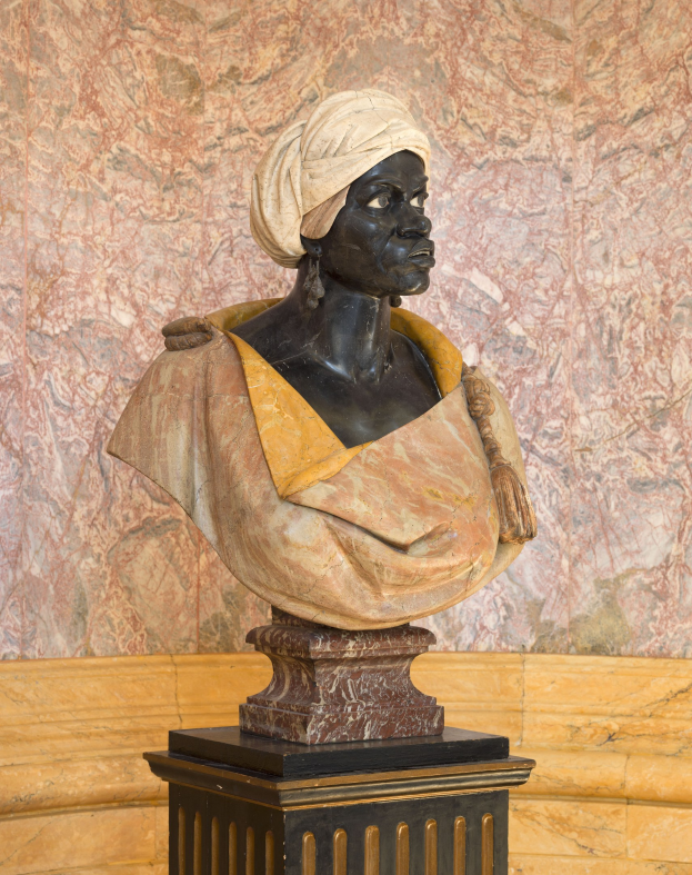 Büste einer Frau mit Turban, die auf einem Sockel vor einer Wand steht, mit Text unten, der besagt, dass sie als erste afrikanische Frau gewählt wurde.