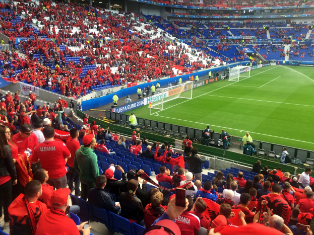 Eine große Menschenmenge in einem Stadion schaut ein Fußballspiel zu, mit Menschen, die auf Stühlen sitzen und stehen, Schildern, einem Torpfosten und Deckenleuchten.