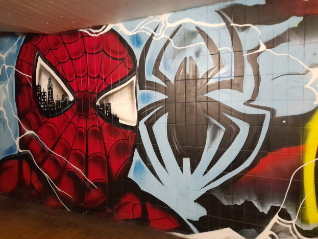 Buntes Wandgemälde von Spider-Man in rot und blauem Kostüm an einer Gebäudewand, mit einem großen Spider-Logo und Webmuster, seine weit aufgerissenen Augen und leicht geöffneten Mund deuten auf Wachsamkeit hin.