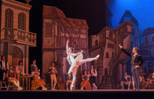 Zwei Tänzer performen auf einer Bühne im Royal Opera House in London, mit einem Publikum im Vordergrund, das auf Stühlen sitzt und steht, und einem Bildschirm im Hintergrund, der Gebäude zeigt.